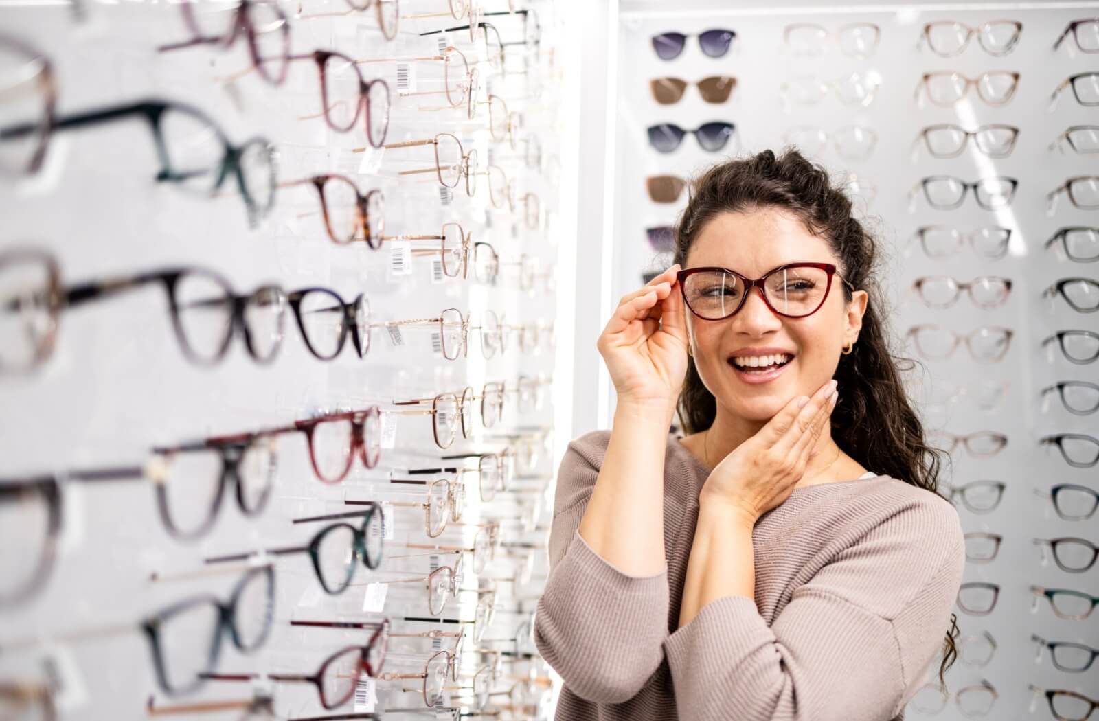 A person smiling while trying on a pair of burgundy cat-eye glasses in an eyewear boutique.