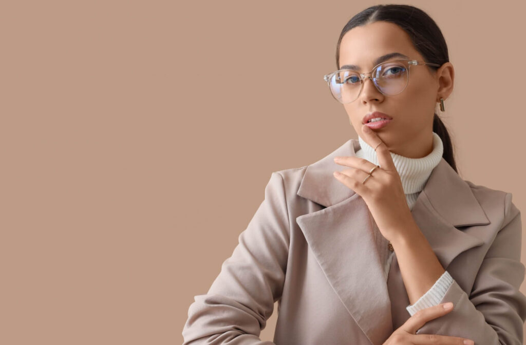A person wearing clear-rimmed glasses and a beige blazer posing against a solid tan background.