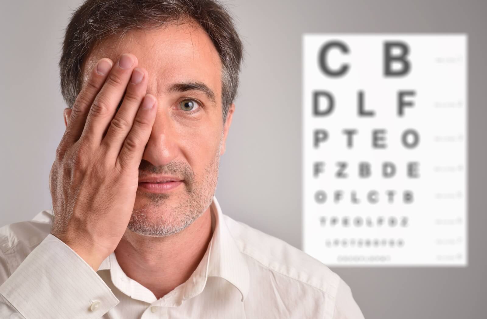 A middle-aged man covering one eye with his hand during a vision test with a blurred eye chart in the background.