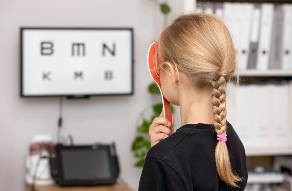 A young girl with a blonde braid seen from behind holding an eye occluder during a pediatric vision exam with a digital eye chart.